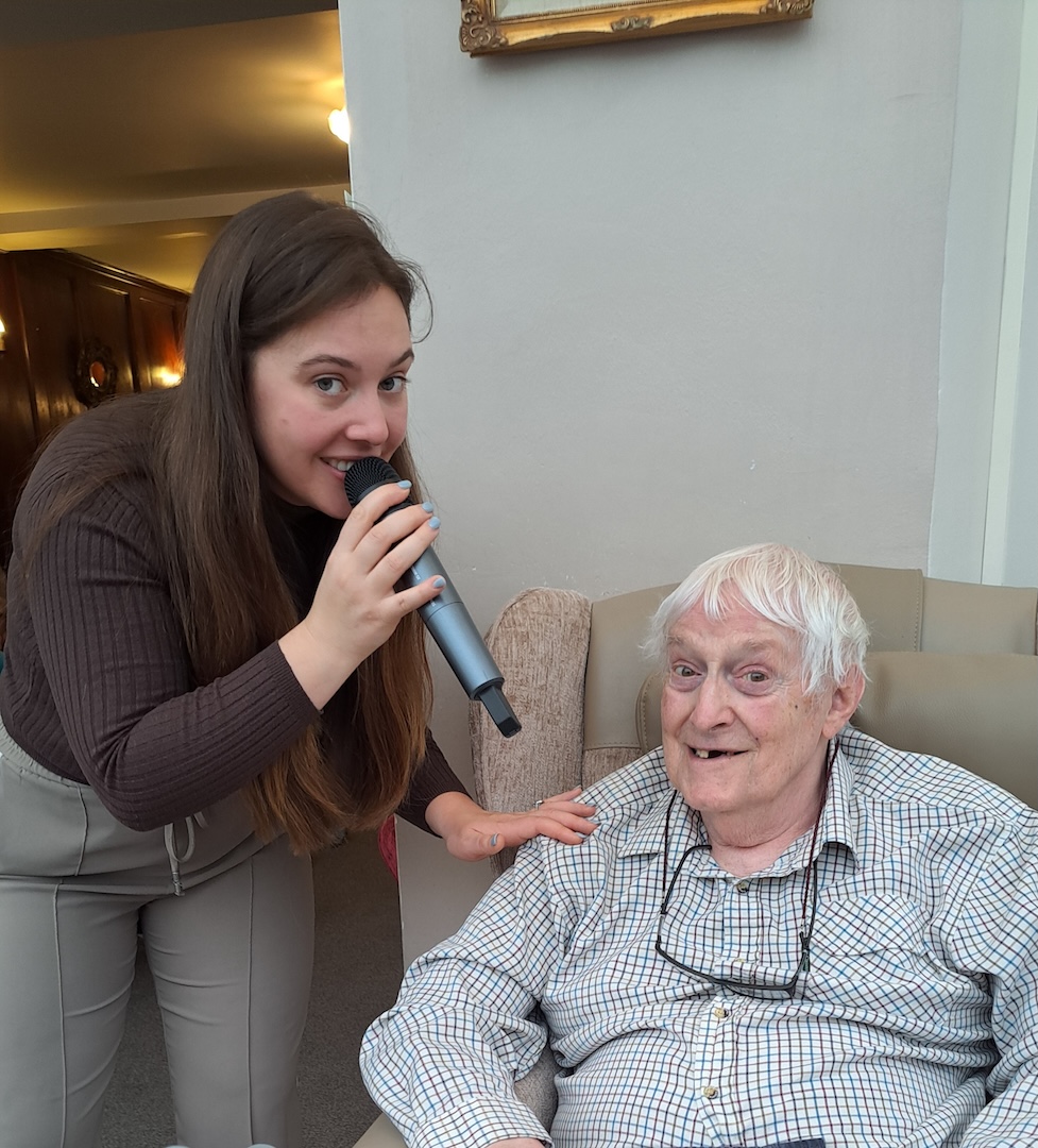 A young women singing into a microphone next to an elderly man