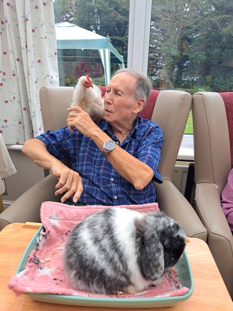 A man with a chicken on his shoulder and a rabbit on a table in front of him