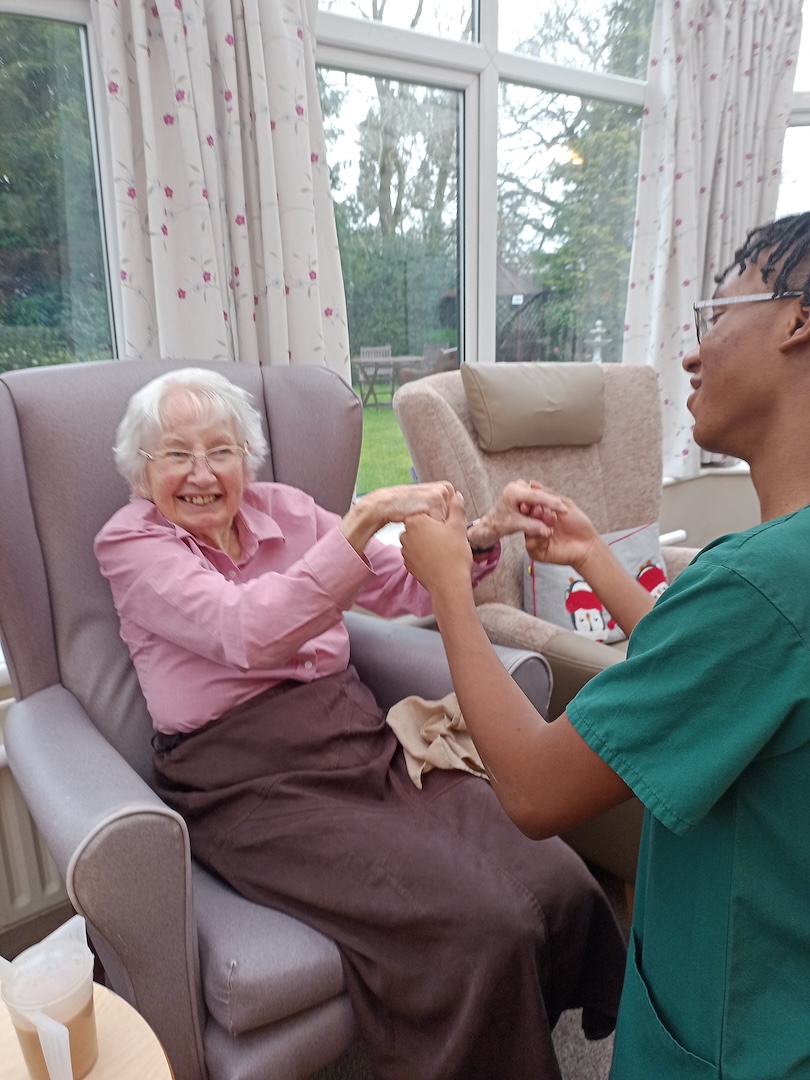 A lady smiling sat in a chair dancing with a member of staff