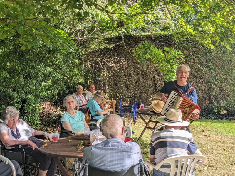 A group of people sat in a garden listening to a man play musical instruments