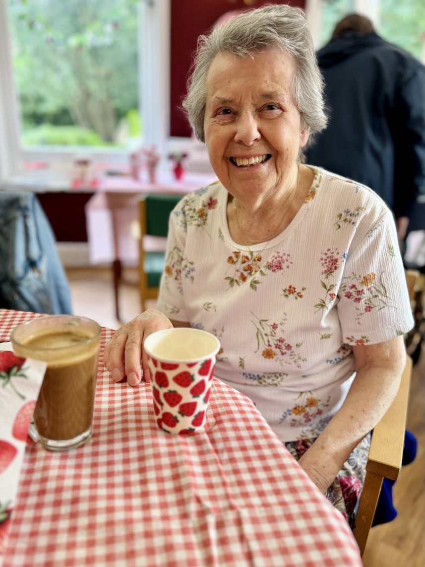 Older woman smiling sat at a table with a drink 