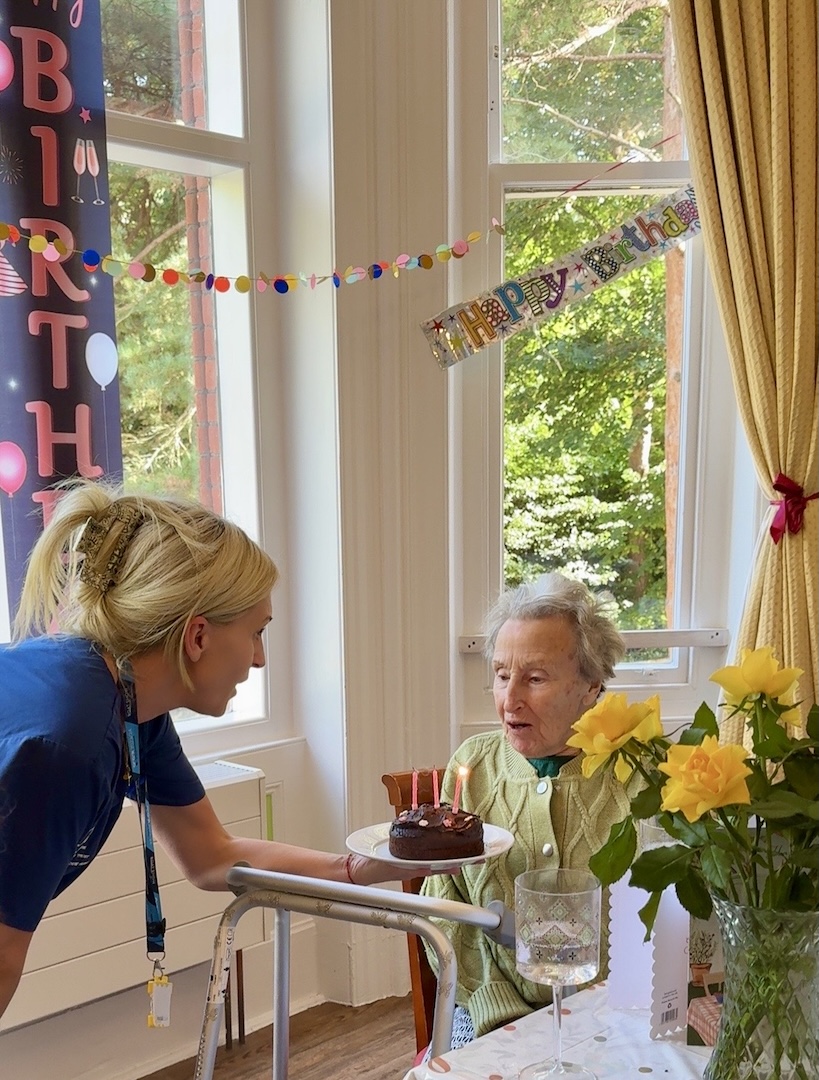 An elderly lady blowing out candles on a birthday cake being presented to her by a staff member