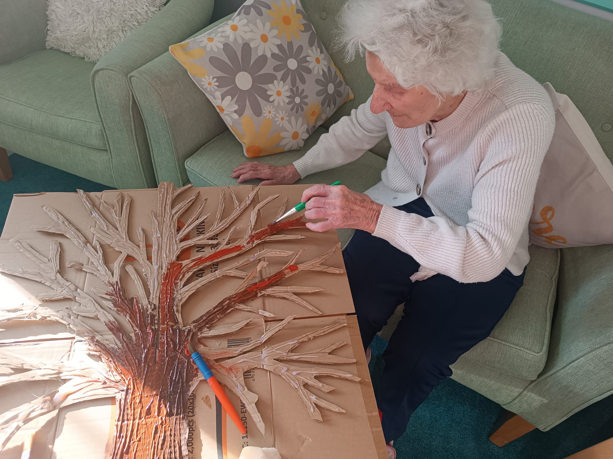 An older lady sat on the sofa working on a painting of a tree