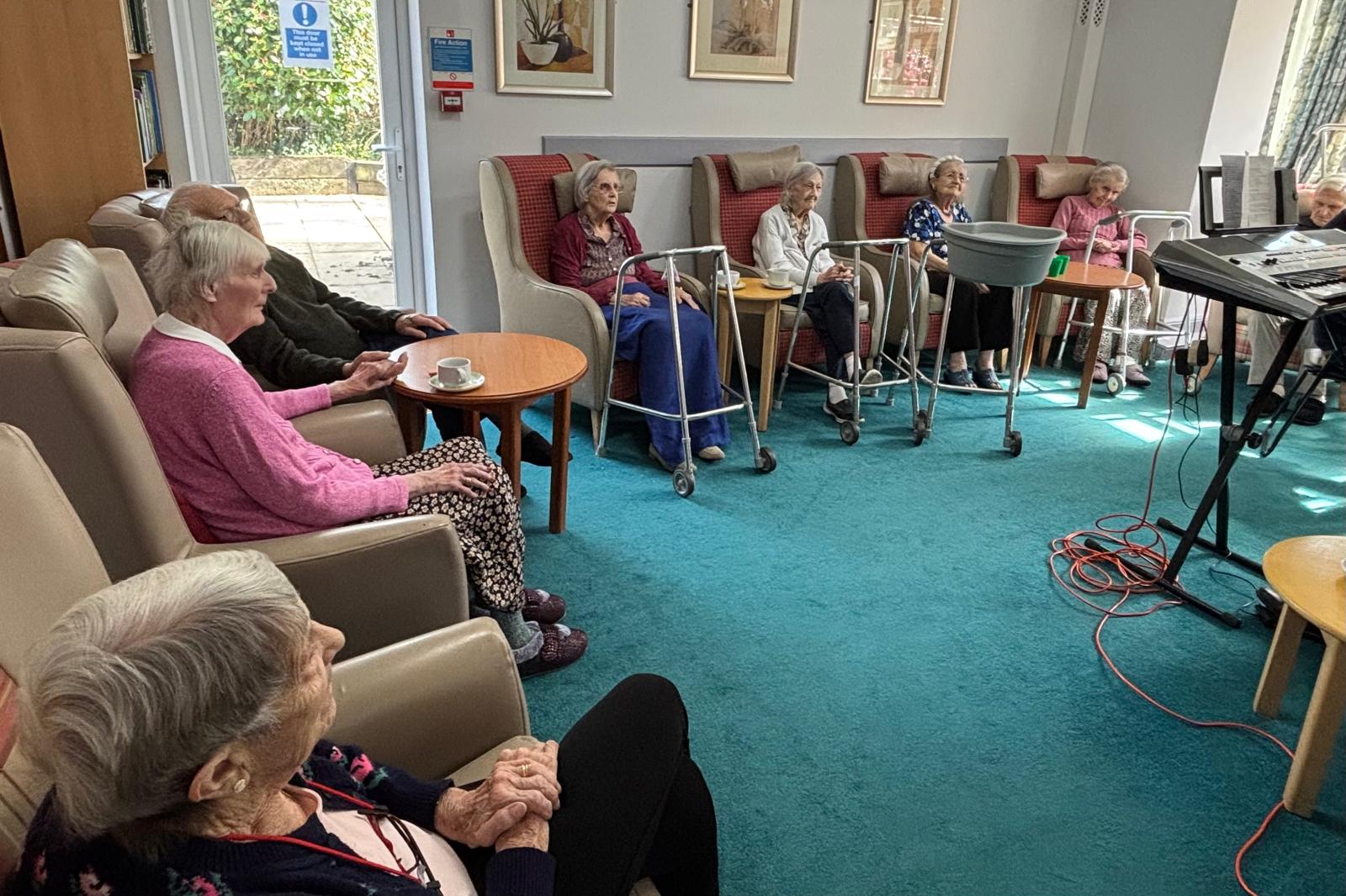A group of elderly people sat in armchairs watching someone play the keyboard