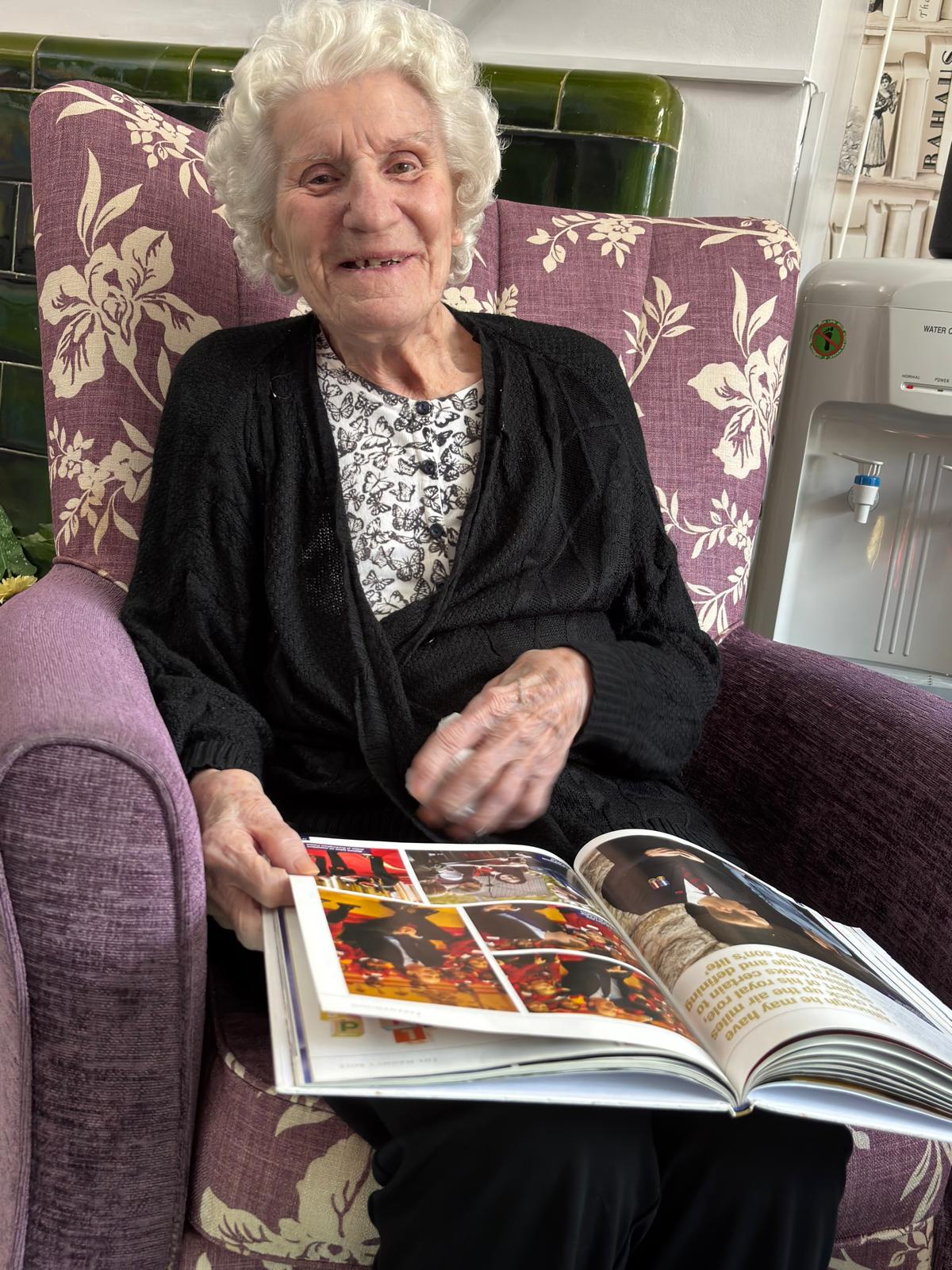 An elderly lady smiling sat in an armchair reading a book