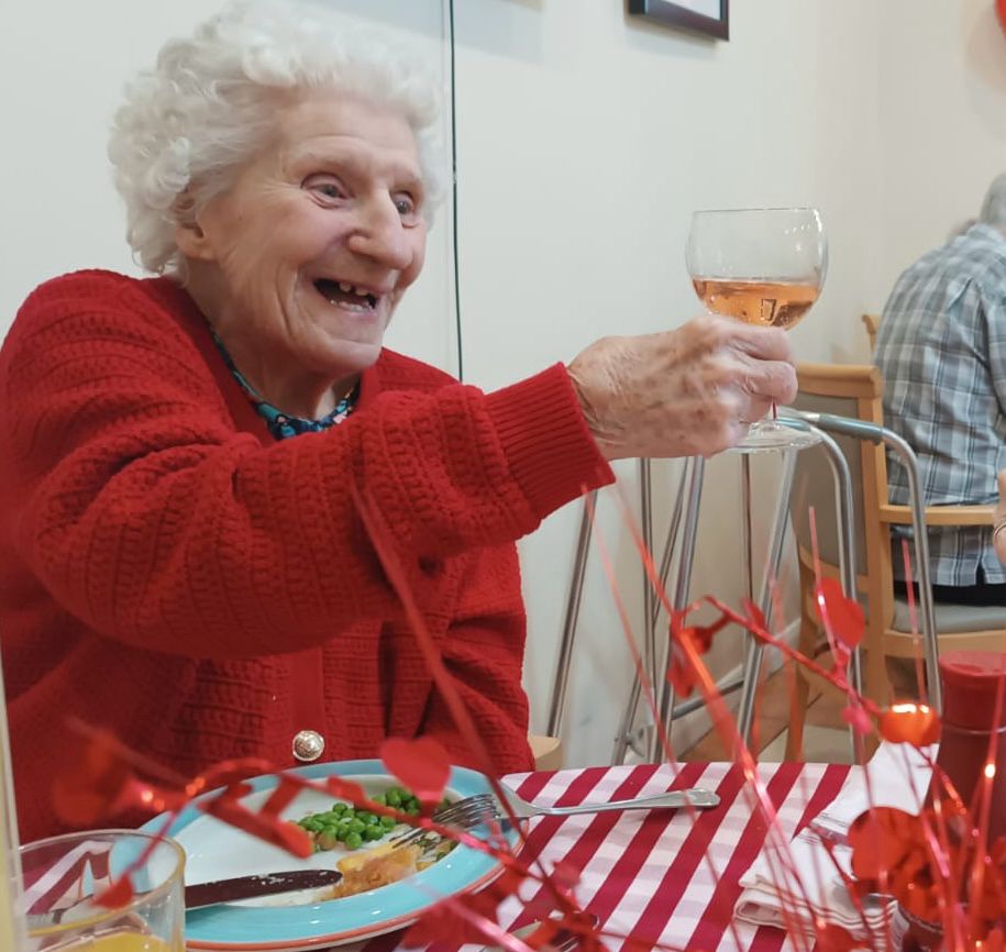 An elderly lady eating dinner saying cheers with a glass of wine