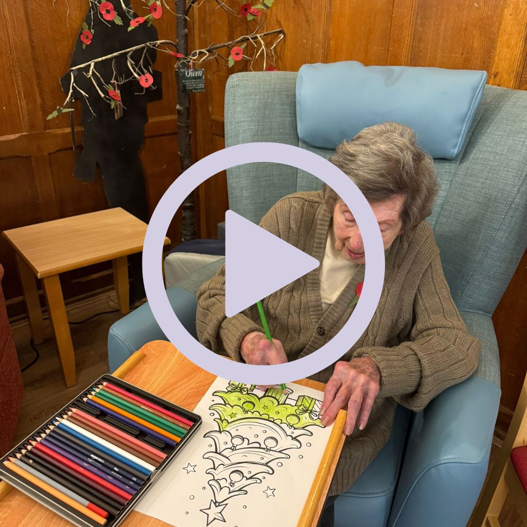 An older woman sat in an armchair colouring in a picture of a Christmas tree