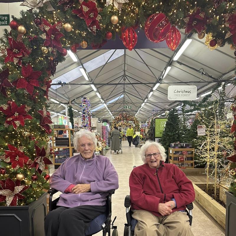Two elderly women in wheelchairs in a garden centre under a festive archway