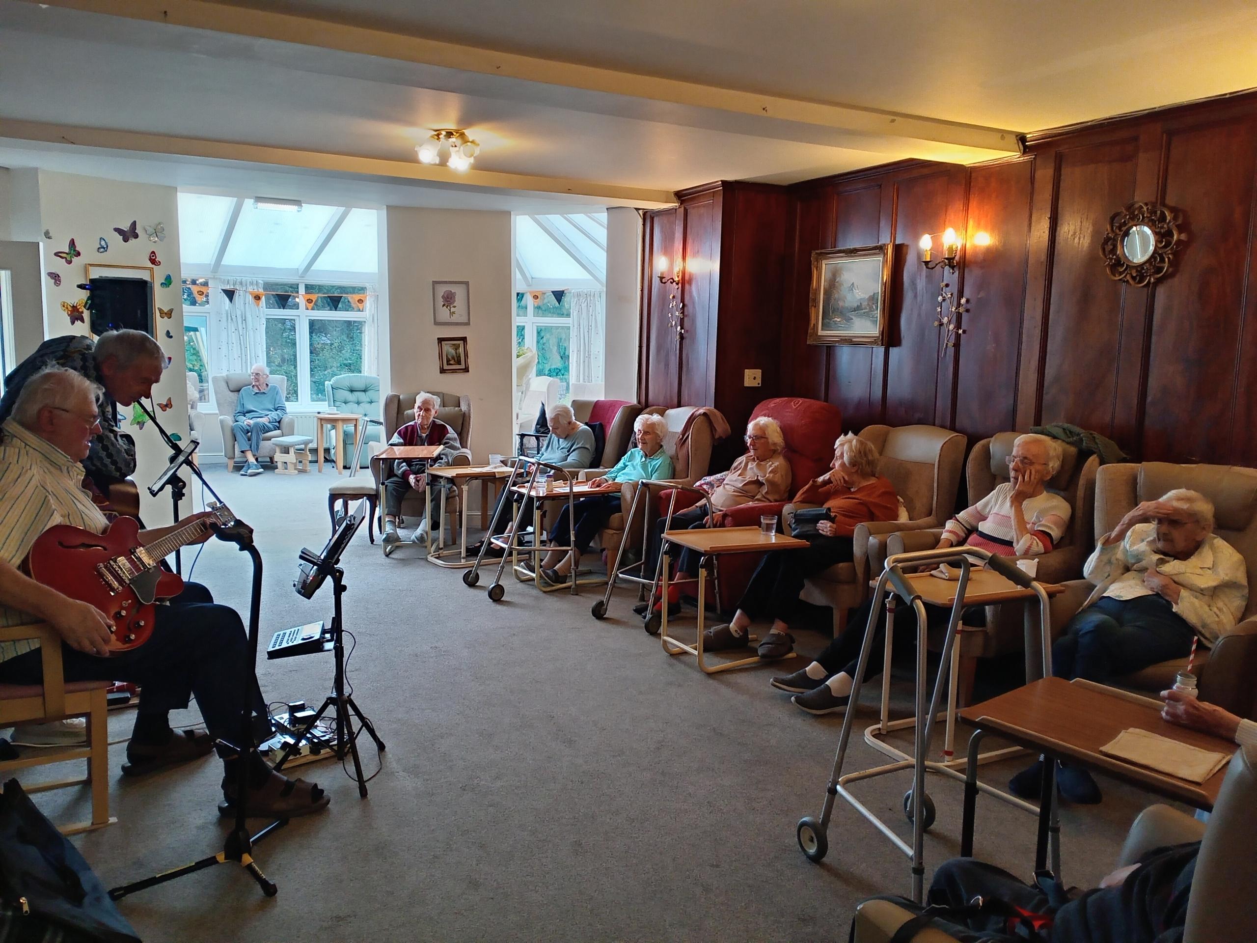 A group of people sat in armchairs watching musical performers playing guitars