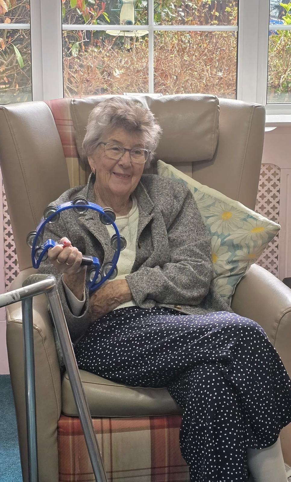 An elderly lady sat in an armchair smiling and holding a tambourine