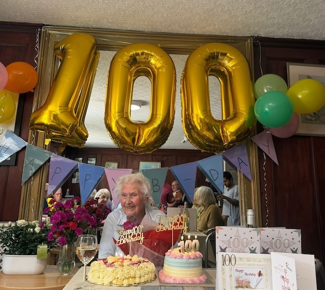 An older woman sat at a table smiling with birthday cakes and balloons.
