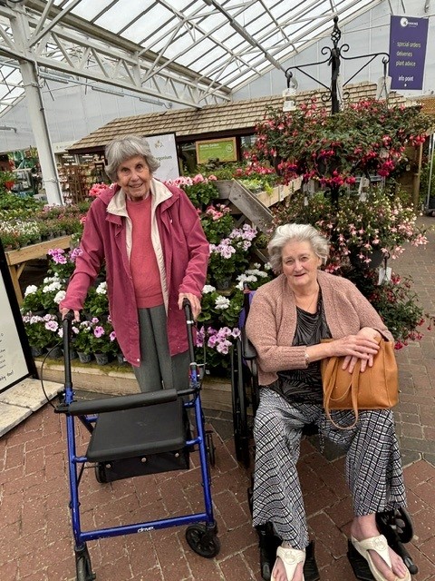 An elderly woman stood in front of flowers at a garden centre with another woman in a wheelchair