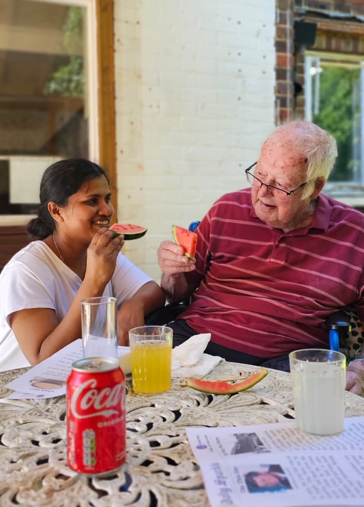 A staff member and resident eating slices on melon sat at a garden table. 