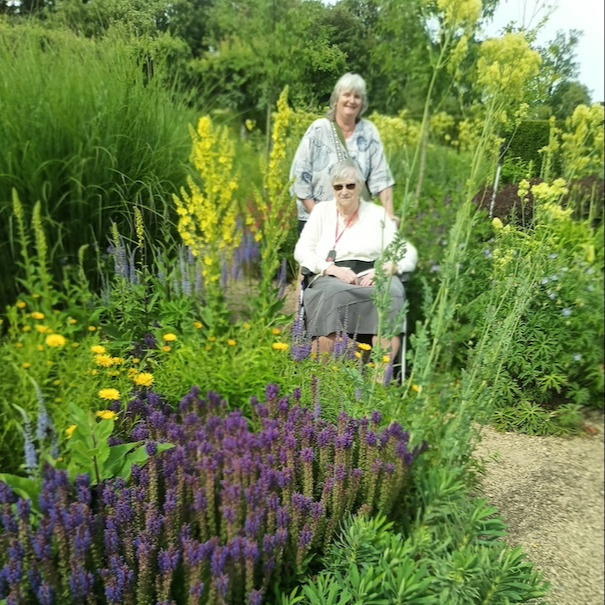 Two people, one in a wheelchair, in a garden full of flowers and shrubbery