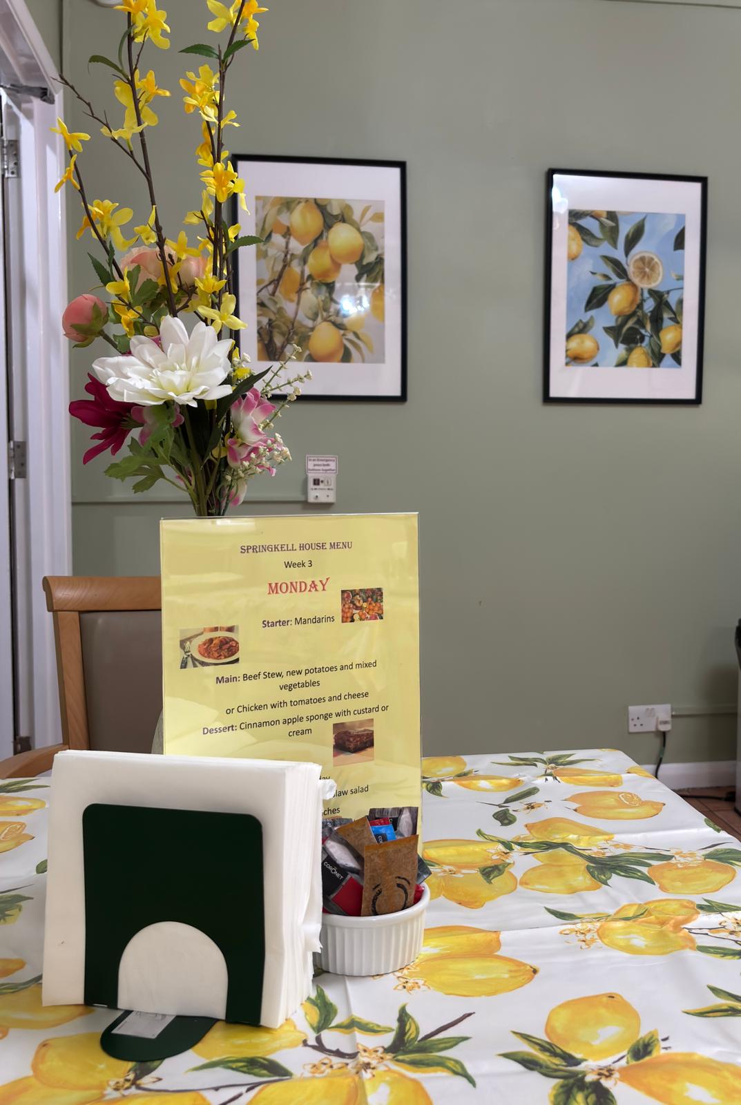 A dining table with flowers and a lemon tabecloth in a dining room