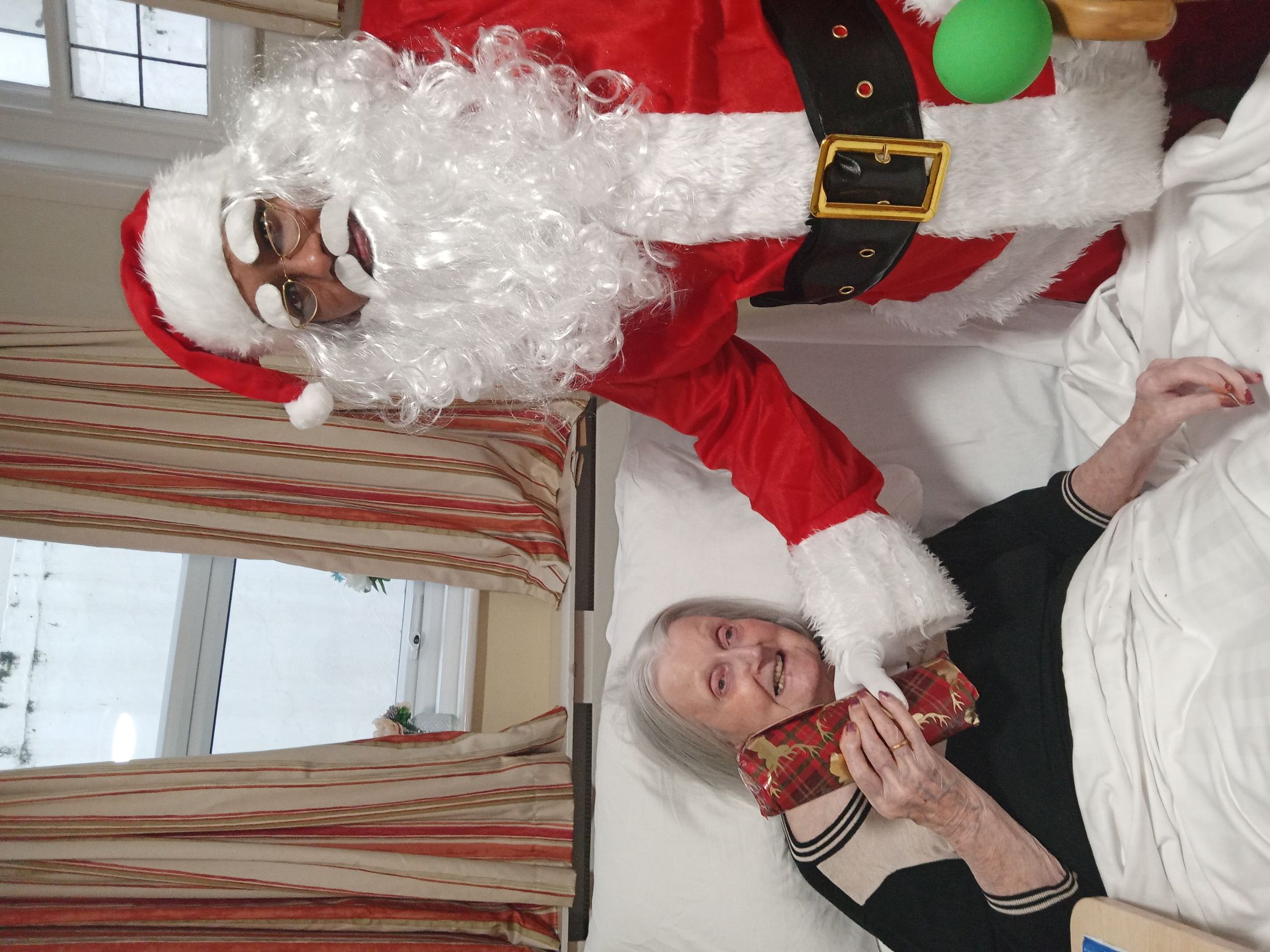 An elderly woman in bed being a gift by a man dressed as santa