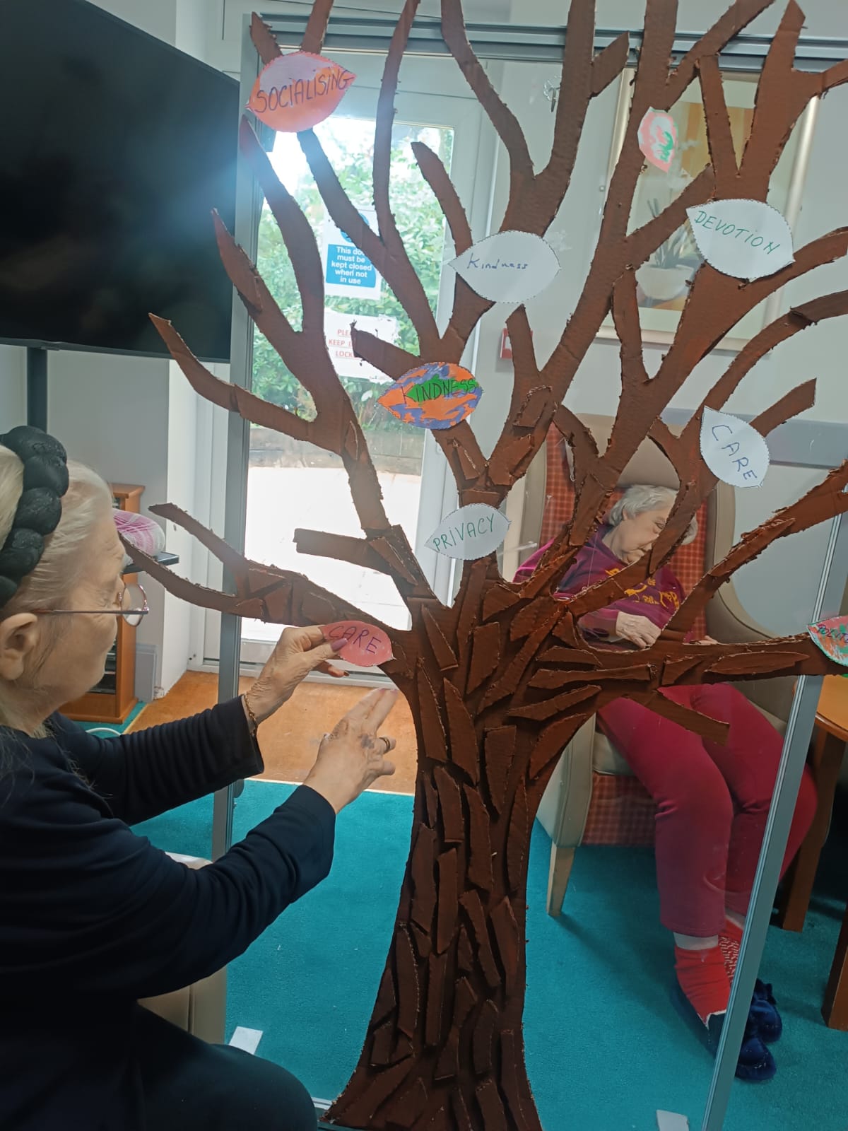 An elderly woman attaching a piece of paper to the branch of an artificial tree. 