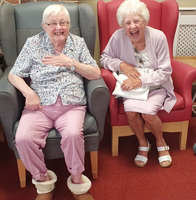 Two laughing elderly women sat in comfortable chairs