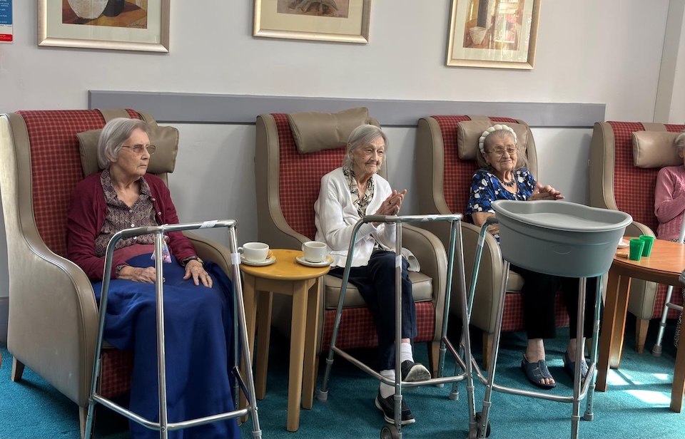 Elderly women sat in armchairs watching some form of entertainment and clapping