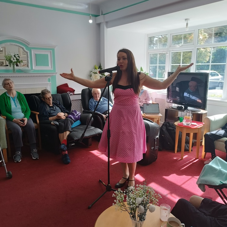 A lady in a pink dress singing to a group of people sat in a lounge