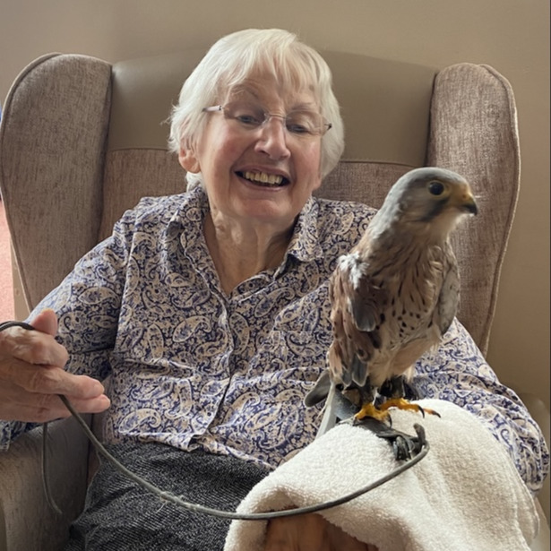 An older woman sat with a bird on her arm and smiling