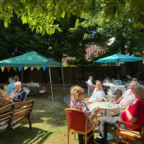 People at a garden party sat on garden furniture in the sushine with gazebos strung with bunting