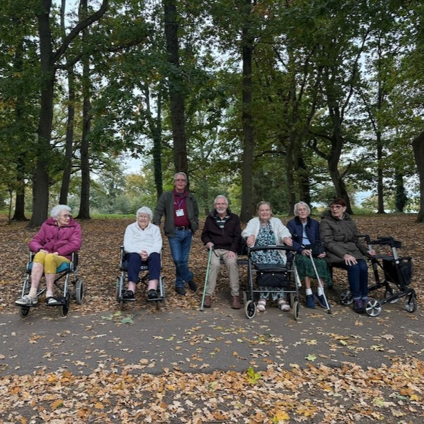 A group of elderly people some in wheelchairs in a woodland going for a walk