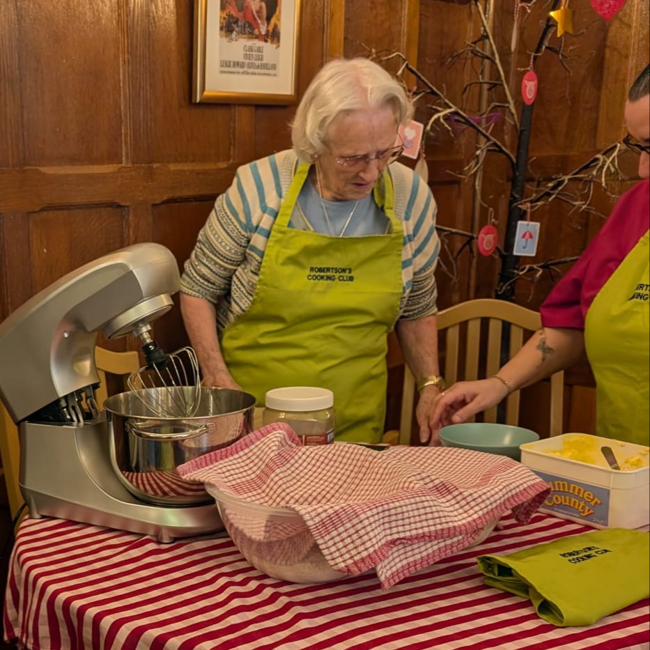 An older woman baking a cake using a stand mixer wearing a green apron being helped by a staff member
