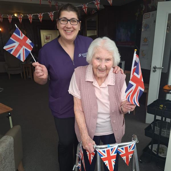 An older woman using a walker with a staff member waving union jack flags at a party