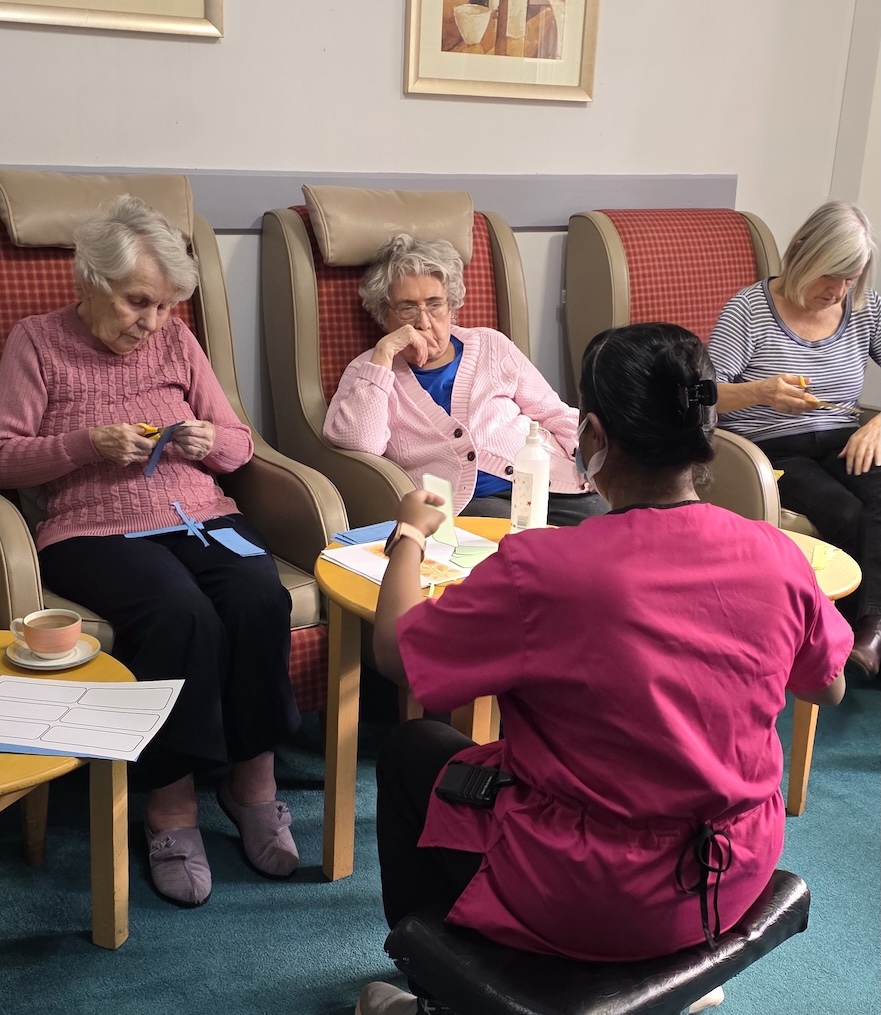 A group of older ladies sat in armchairs taking part in an activity run by a member of staff sat on the floor in front of them.