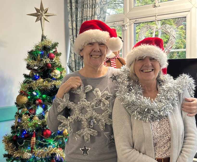 Two ladies stood in front of a Christmas tree wearing Christmas hats and holding decorations.