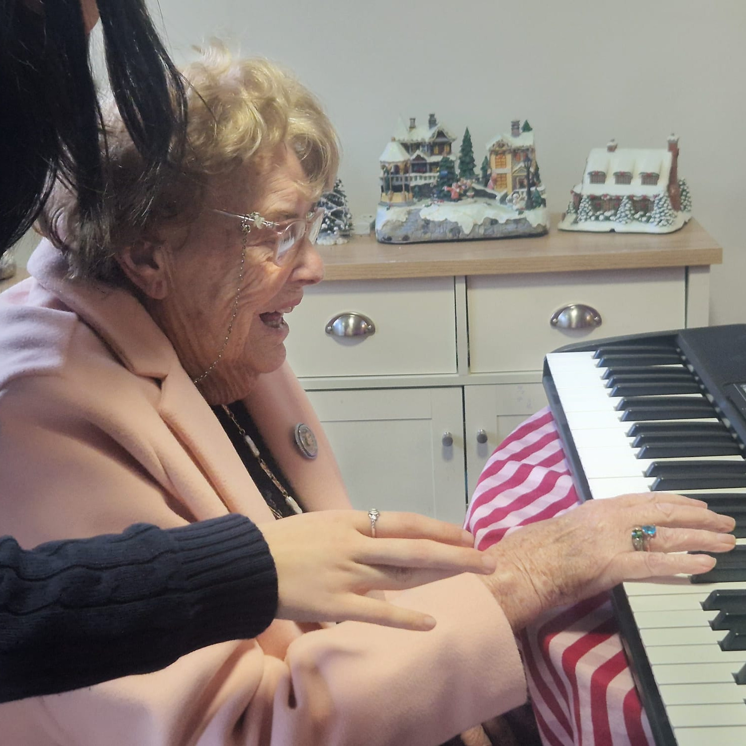 An older lady playing the piano being helped by a young volunteer