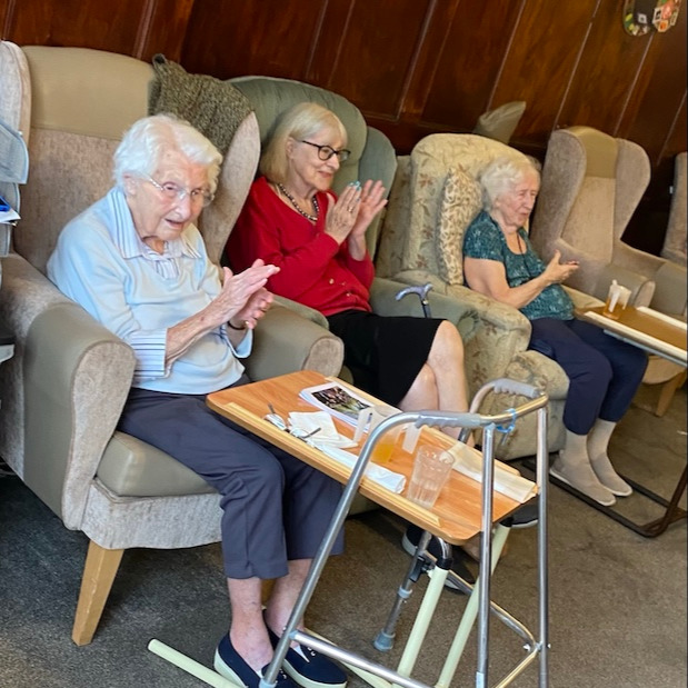 Three older ladies sat on armchairs clapping as they listen to music