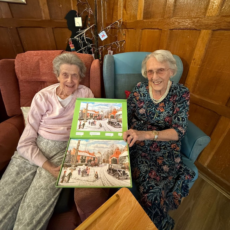 Two older ladies sat together smilling holdig a completed puzzle