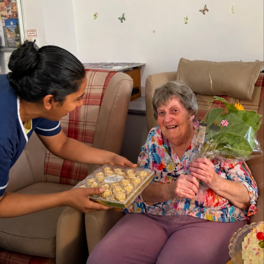 A staff member presenting chocolates to an older woman holding flowers