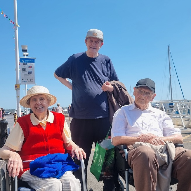 A group of people at the quay in the sunshine with boats in the background