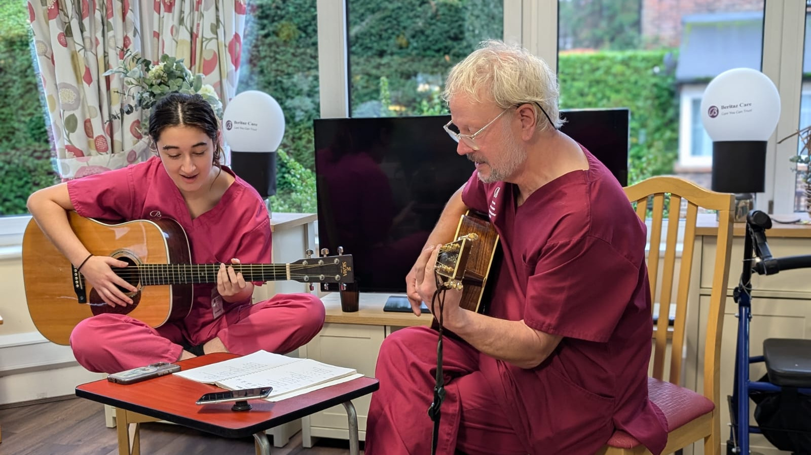 Two people playing guitars sat on wooden chairs. 
