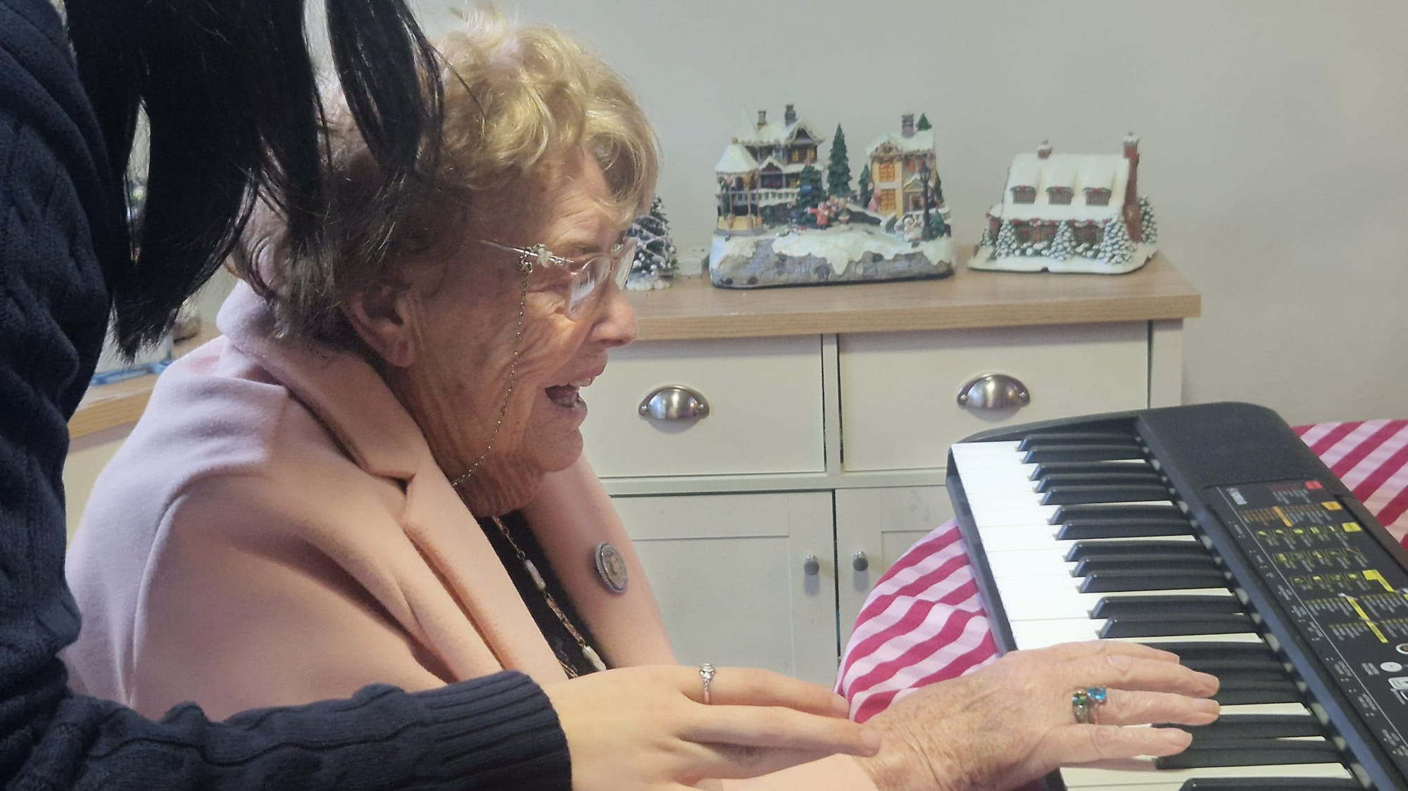 An older woman playing the keyboard with another person stood behind helping
