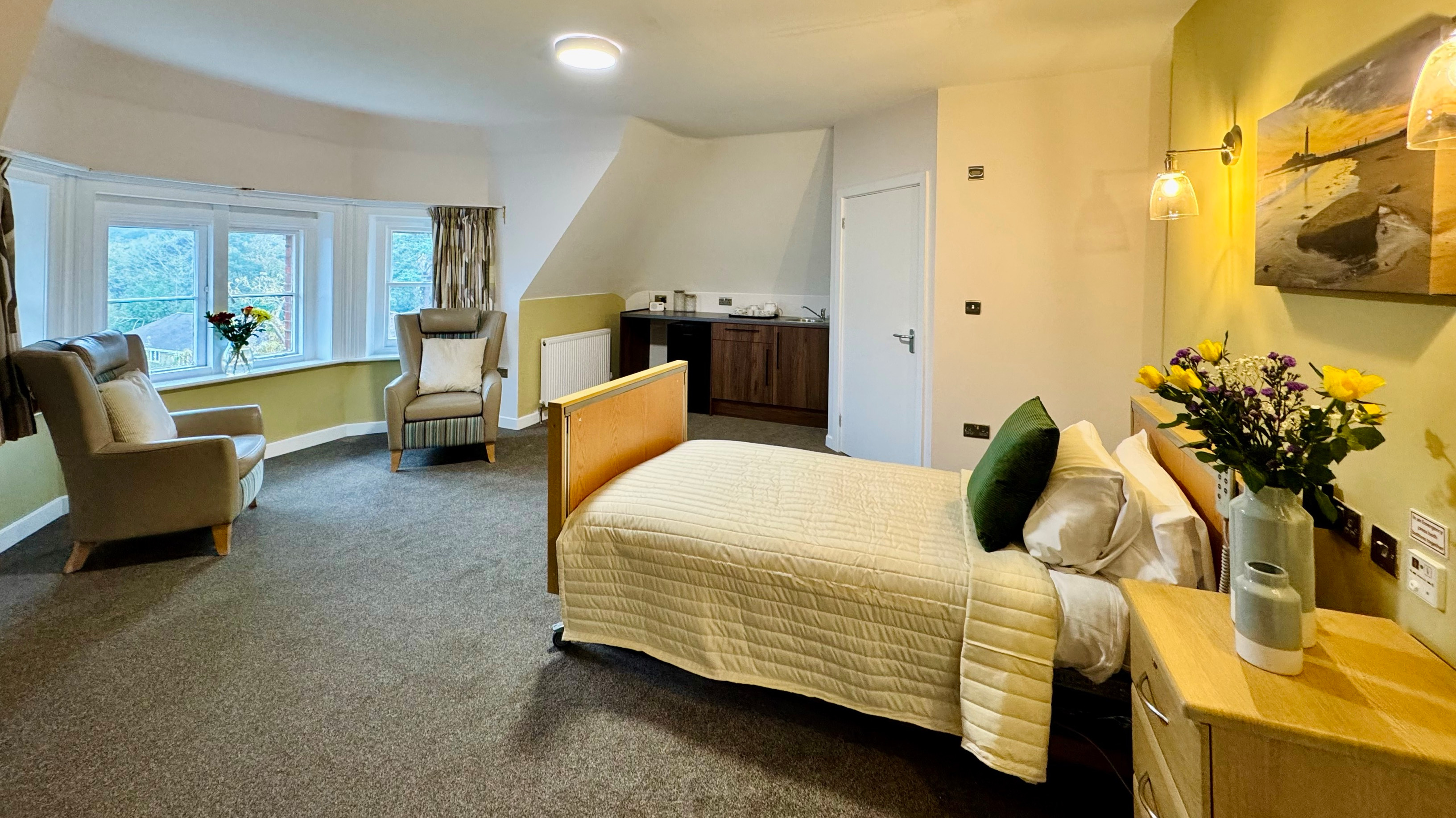 A clean room with a comfortable bed and two chairs, flowers are on the bedside table