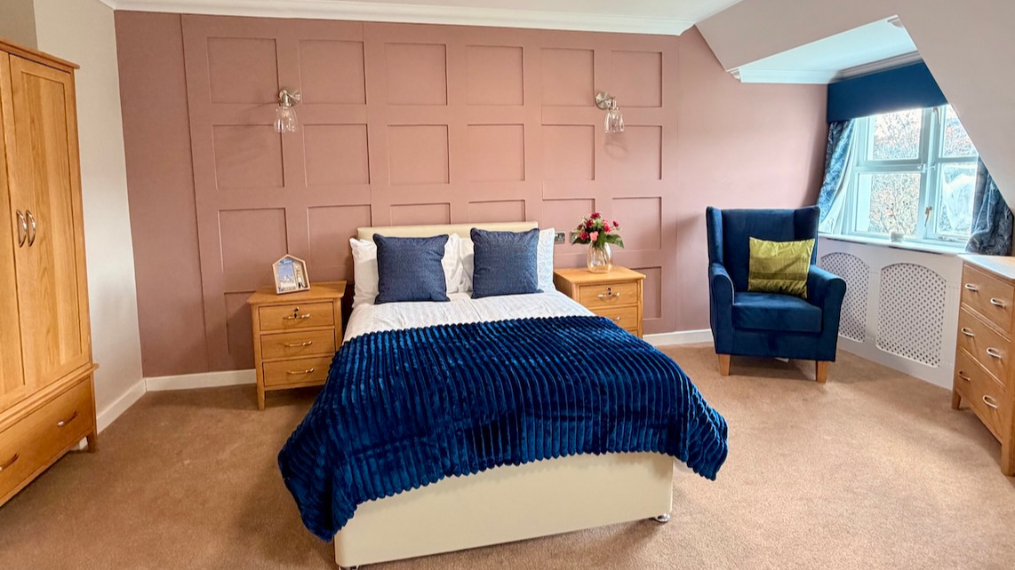 A bright well decorated room with a comfortable bed and chair, flowers are on the bedside table
